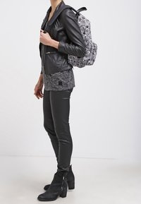 Giubbotto di pelle, top grigio texturizzato, leggings neri e stivali alla caviglia; zaino a righe zebrate sulla spalla; vista laterale, sfondo neutro.
