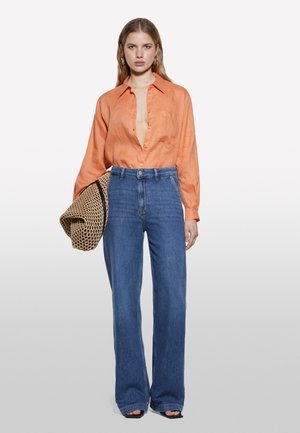 Chemise en lin orange avec un décolleté en V prononcé, associée à un pantalon en denim bleu taille haute évasé. Le modèle tient un sac tissé. Texture lisse et style décontracté.