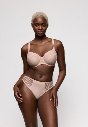 Vrouw met kort blond haar, gekleed in een lichtroze kanten beha en bijpassende slip, staand tegen een effen grijze achtergrond.