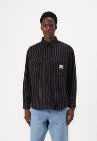Black and white striped button-up shirt with two chest pockets, a collar, and a logo patch, featuring a relaxed fit and long sleeves.