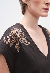 Μαύρο τοπ με λαιμόκοψη σε σχήμα V, διακοσμημένο με κεντητά λουλουδένια σχέδια σε μπεζ και καφέ χρώματα στους ώμους. Το ύφασμα έχει υφή και απαλό φινίρισμα.