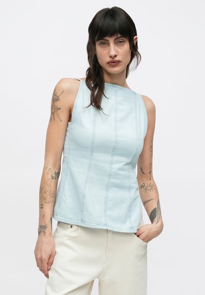 Γυναίκα με τατουάζ και στα δύο χέρια, που φοράει αμάνικη ανοιχτό μπλε μπλούζα και εκρού παντελόνι, στέκεται με το ένα χέρι στην τσέπη.