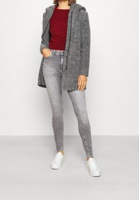 Manteau en chevrons gris avec capuche, associé à un pull bordeaux et à un jean skinny gris clair. Des baskets blanches complètent la tenue.