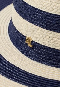 Καπέλο από ψάθα με εναλλασσόμενες λωρίδες σε ναυτικό μπλε και μπεζ, με υφή και ένα χρυσό μεταλλικό λογότυπο ως διακοσμητικό στοιχείο.