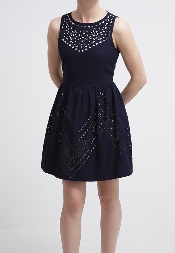 Femme portant une robe bleu marine sans manches avec des motifs découpés géométriques sur le haut de la poitrine et la jupe, debout les mains derrière le dos.