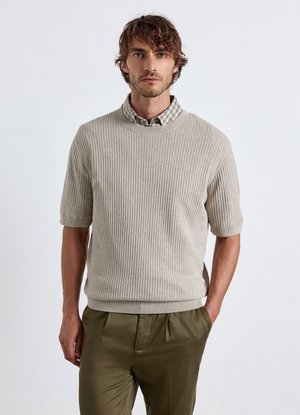 Mann trägt einen beige gerippten Kurzarmpullover über einem karierten Hemd und olivgrünen Hosen, steht mit einer Hand in der Tasche vor weißem Hintergrund.