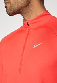 Κόκκινη μπλούζα Nike με φερμουάρ τεταρτημόριο, κατασκευασμένη από ελαστικό ύφασμα, με ασημένιο λογότυπο στο στήθος και σχεδίαση με ψηλό γιακά. Λεία υφή.