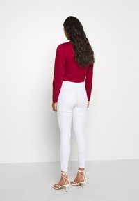Topa roja de manga larga combinada con jeans ajustados blancos. La parte superior tiene hombros abullonados; los jeans son entallados con bolsillos traseros y un diseño clásico.