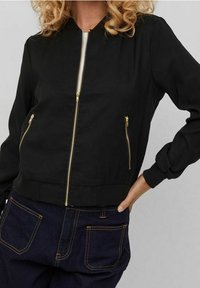 Černá bomber bunda se zlatým zipem, dvěma předními kapsami, žebrovanými manžetami a přiléhavým pasem. Hladká tkanina a jednoduchý design.