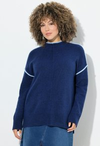 Frau mit lockigem Haar, die silberne Ohrringe, einen blauen Pullover mit hellblauen Akzenten und einen Jeansrock trägt und vor einem weißen Hintergrund steht.