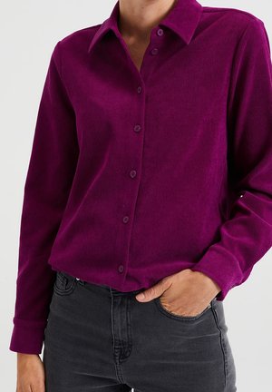 Vrouw draagt een paarse corduroy blouse met lange mouwen, ingestopt in een zwarte spijkerbroek, met één hand in de zak.