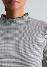 Vrouw met een zwart-wit gestreept topje met gerafelde halslijn, zichtbaar van schouders tot lippen tegen een effen lichte achtergrond.