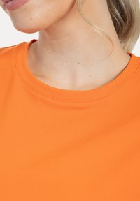 Oranje top met ronde hals van gladde stof, met een zachte textuur en nette stiksels langs de halslijn. Close-up weergave.