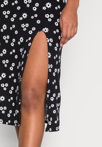 Černé šaty s bílým květinovým vzorem, s výstřihem až k stehnům a hladkou texturou, předvádějící přiléhavý siluetu a moderní design.