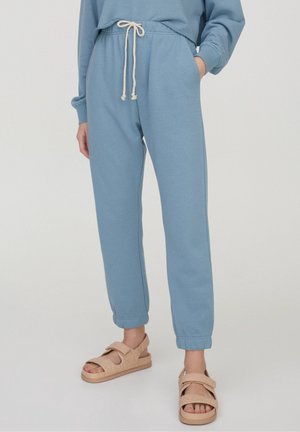 Personne portant un pantalon de survêtement bleu clair avec cordon de serrage et des sandales beiges à bout ouvert, debout sur un fond uni clair.
