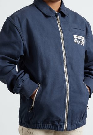 Marineblauwe ritsjack gemaakt van stevig materiaal, met een kraaghals, ribgebreide manchetten en een 'All Star' logo-embleem op de borst.