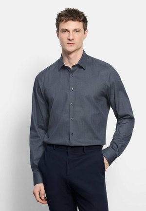 Mann trägt ein dunkel gemustertes Hemd mit Knöpfen und marineblaue Hose, steht mit einer Hand in der Tasche vor einem weißen Hintergrund.