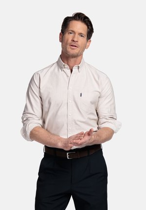 Man in een wit overhemd met opgerolde mouwen en donkere broek staat met gevouwen handen en kijkt vooruit tegen een effen witte achtergrond.