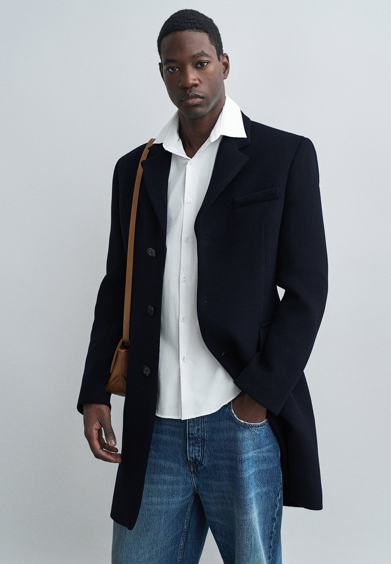 Navy wool coat with notch lapel and four buttons, worn over a white shirt. Pairing with light blue denim jeans and a brown shoulder bag.