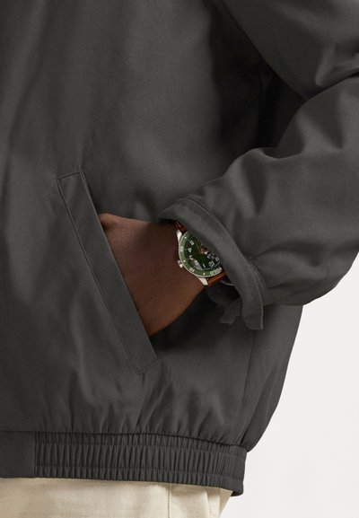 Chaqueta de cremallera de color gris oscuro con dobladillo elástico, que presenta un bolsillo lateral y un puño donde se muestra un reloj verde con correa de cuero marrón.