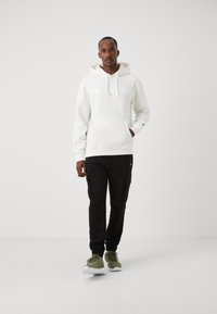 Felpa bianca con cappuccio, tasca anteriore e logo ricamato, abbinata a pantaloni cargo neri e scarpe da ginnastica verdi.