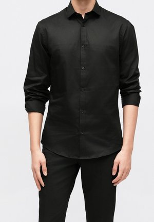 Homme portant une chemise noire à manches longues boutonnée, manches retroussées, et un pantalon noir devant un fond uni.