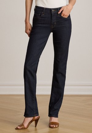 Le modèle porte un jean foncé à jambes droites, un haut blanc et des sandales marron à talons, une main dans une poche avant.