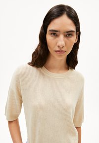 Beige geripptes Strickpullover mit kurzen Ärmeln und rundem Halsausschnitt. Weiche Textur und subtiler Glanz. Betont durch feine vertikale Linien.