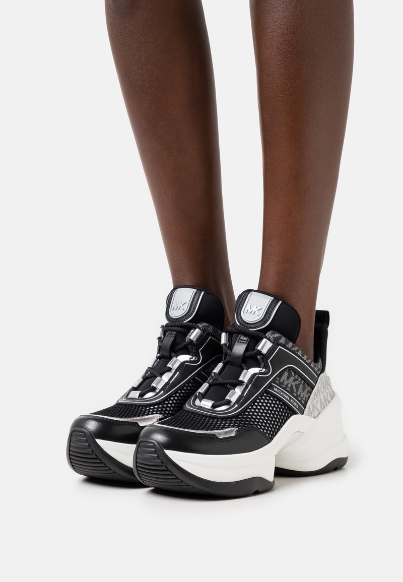 Baskets de sport noires avec une tige en mesh, une semelle blanche prononcée, des accents argentés et des détails texturés au niveau du talon.
