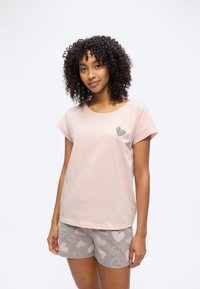 Γυναίκα με σγουρά μαλλιά, που φοράει ένα ανοιχτό ροζ κοντομάνικο πουκάμισο και γκρι σορτς με ροζ σχέδια καρδιών, στέκεται μπροστά σε λευκό φόντο.