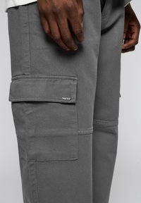 Grijze cargobroek van stevig materiaal, met zijzakken, gestikte accentlijnen en een metalen logo label. Close-up van de hand op het been.