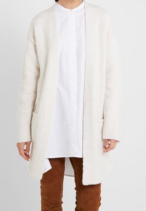 Person iført en lang, off-white strikket cardigan over en hvid skjorte med knapper og brune bukser, stående foran en lys baggrund.