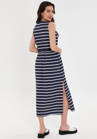 Marineblau-weiß gestreifte Maxikleid mit tailliertem Schnitt und seitlichem Schlitz; ärmelloses Design; weiches Material mit leichtem Glanz; getragen mit weißen Slides.