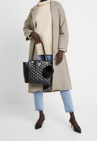 Sac à main matelassé noir avec ferrures argentées et un pompon en fourrure, porté par une personne vêtue d'un pull beige et d'un manteau gris, avec un jean bleu.
