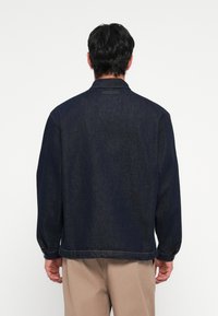 Temno modra denim jakna z dolgimi rokavi in gumbi na manšeti. Ima klasičen ovratnik in ravno rob. Ni vidnih oznak blagovne znamke.