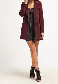 Burgundy zip-up jacka med långa ärmar, sidofickor och skräddarsydd passform; kombinerat med en denimklänning och svarta hälinspirerade sandaler.