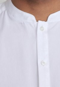 Camisa blanca abotonada con cuello mandarín, textura suave y un ligero brillo. Se observan dos botones en la parte frontal.