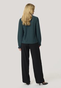 Teal gemusterte Langarmbluse mit elastischen Bündchen, kombiniert mit weiten schwarzen Hosen und Absatzschuhen, von hinten betrachtet.