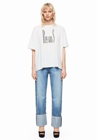 T-shirt bianco oversize con grafica "PEPE GIRLS", abbinato a jeans a gamba larga di colore blu chiaro con orli arrotolati e scarpe platform nere.
