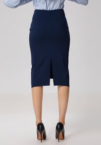 Jupe crayon navy avec fente arrière, coupe ajustée et fermeture éclair dissimulée. Portée avec des chaussures à talons noirs. Texture de tissu lisse.