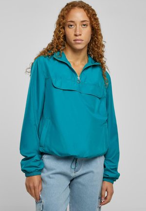 Teal vindjakke med høy krage, frontglidelås og stor lomme. Har elastiske mansjetter og snor i bunnen. Glatt stofftekstur.