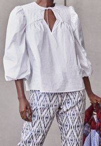 Blusa di cotone bianca con maniche a palloncino e scollatura a buco della serratura, abbinata a pantaloni con motivo geometrico blu e bianco.