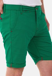 Grüne Baumwollshorts mit aufgerolltem Saum, roten Akzenten, Reißverschluss vorne und seitlichen Taschen. Das Material hat einen glatten, strukturierten Look.