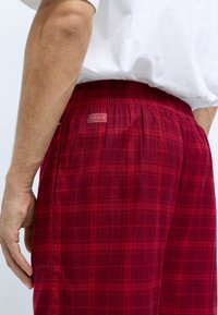 Pantalons de détente à carreaux rouges avec une taille élastique et des poches latérales. Présente une petite étiquette de marque sur l'arrière de la taille.