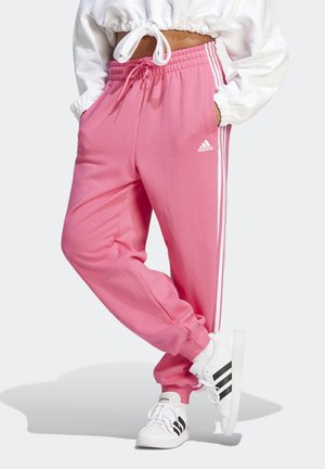 Persoon die roze Adidas trainingsbroek draagt met witte drie strepen en witte sneakers met zwarte strepen, staand met handen in de zakken.