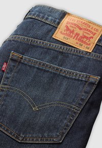 Blugi Levi's de culoare albastru închis, pliați, arătând buzunarul din spate cu cusături portocalii, eticheta roșie Levi's și eticheta din piele bej cu logo-ul și detaliile mărimii.