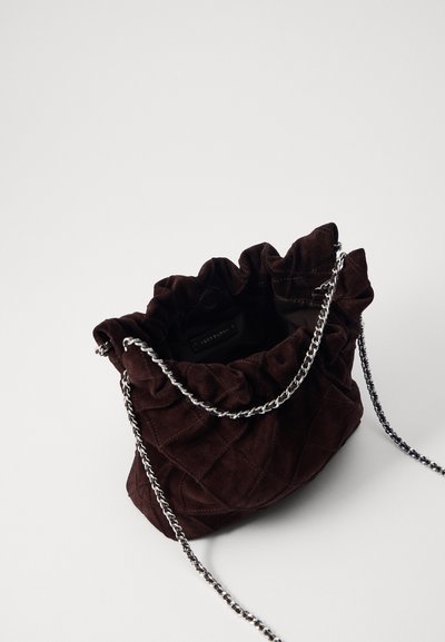 Geantă din suede maro quilted, cu vârf ondulat, curea din lanț argintiu și interior deschis cu etichetă. Suprafață texturată cu un finisaj moale.
