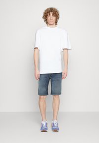 Camiseta de algodón blanca, de corte relajado, combinada con shorts de mezclilla azul. Usada con zapatillas deportivas azul claro y blancas, con suela de goma marrón.