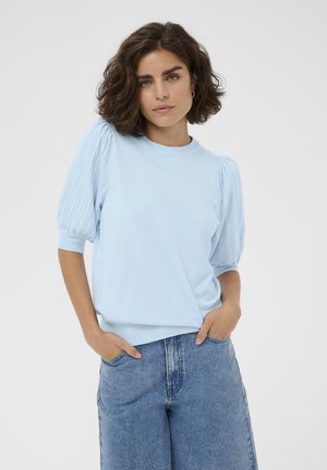 Femme aux cheveux courts et bouclés portant un pull bleu clair à manches courtes bouffantes et un jean denim bleu, les mains dans les poches.