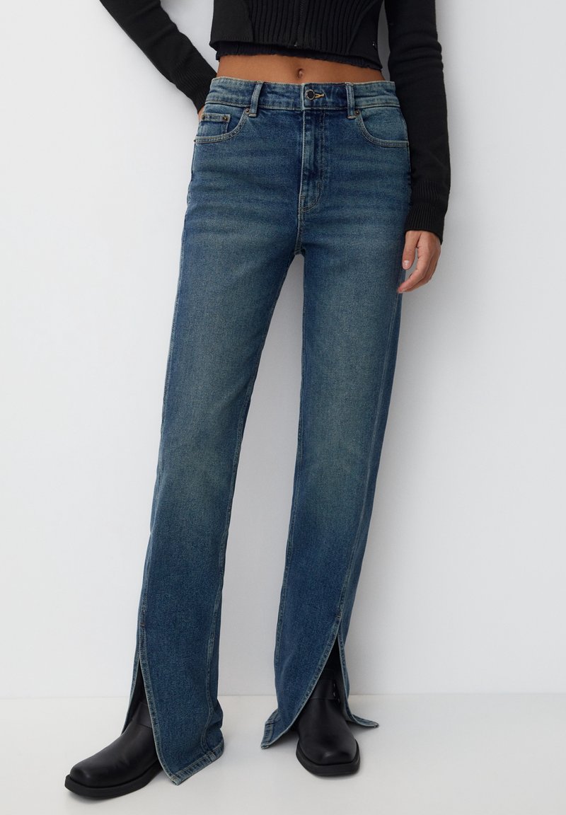 Femme portant un jean bleu taille mi-haute avec des ourlets fendillés à l'avant, assorti de bottines noires et d'un pull court noir, debout contre un mur blanc.